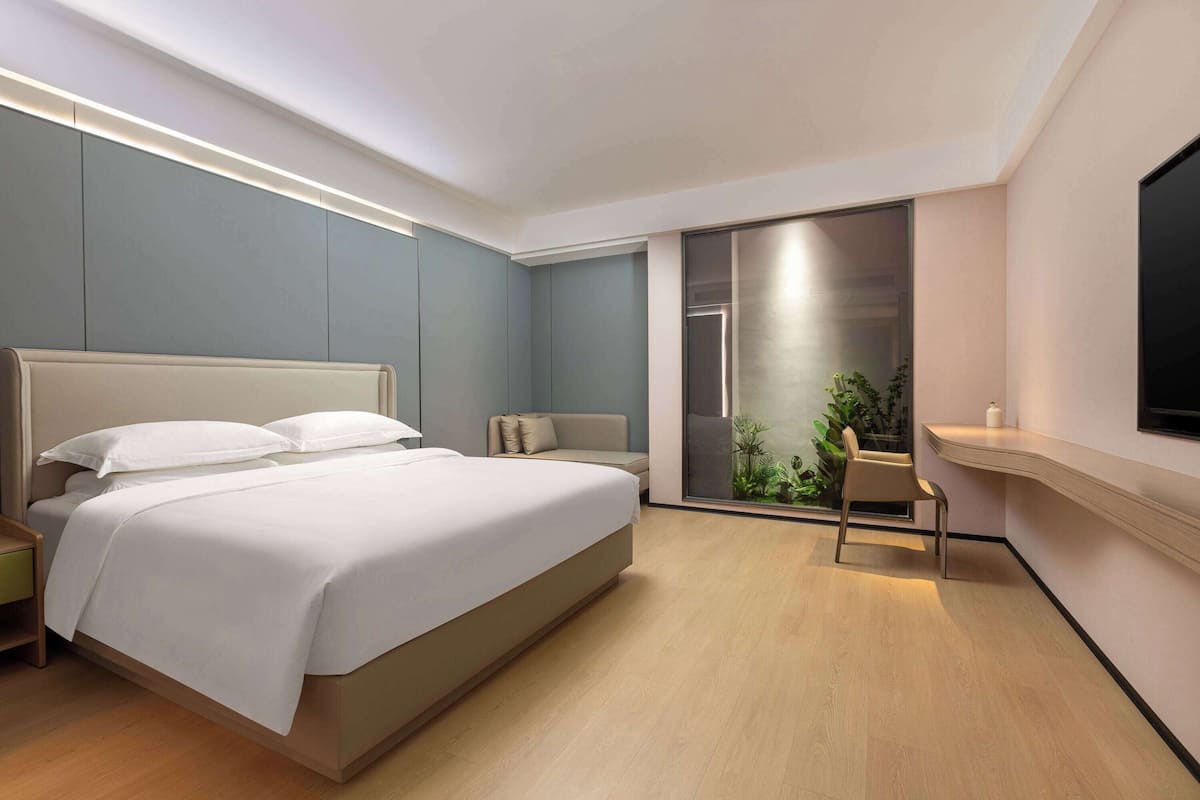 A modern bedroom with a large bed, a desk, and a sliding glass door leading to a balcony with plants.