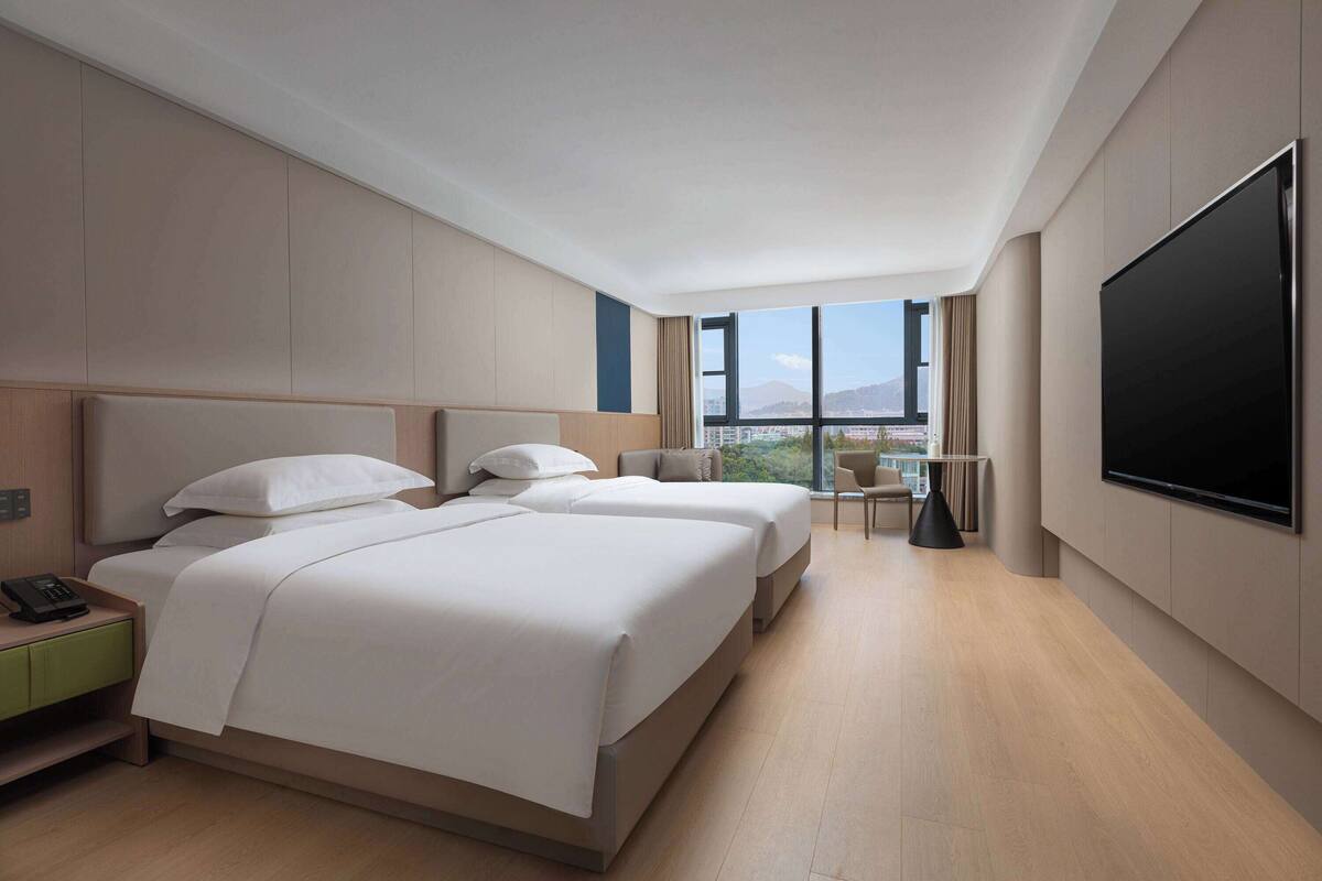 A modern hotel room with two beds, a TV, a desk, and a view of the city.