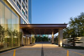 Una entrada de hotel moderna con un paseo techado, fachada de vidrio y zona ajardinada.