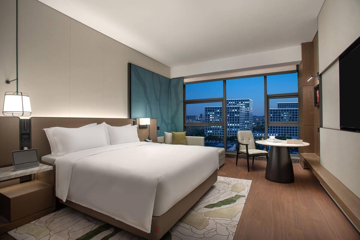 A modern hotel room with a large bed, a desk with a laptop, a round table, and a city view.
