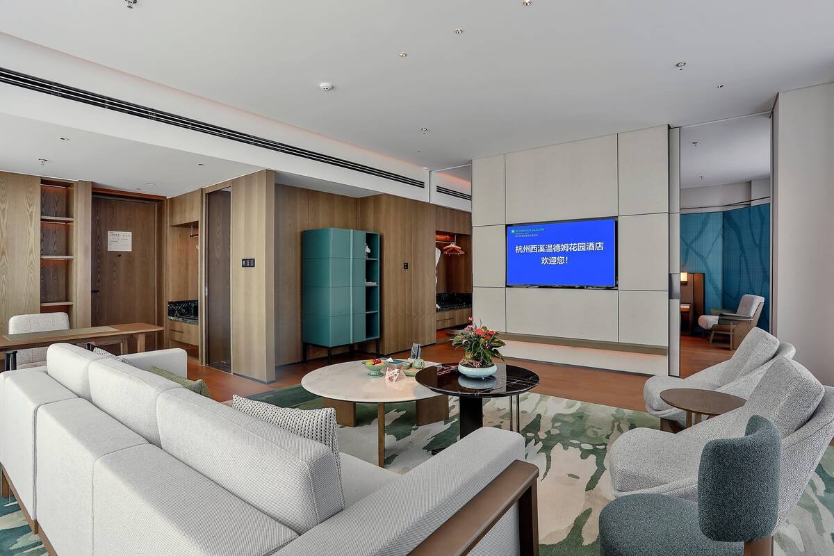 A modern hotel lobby with a sofa, armchairs, a coffee table, a TV screen, and a wooden cabinet.