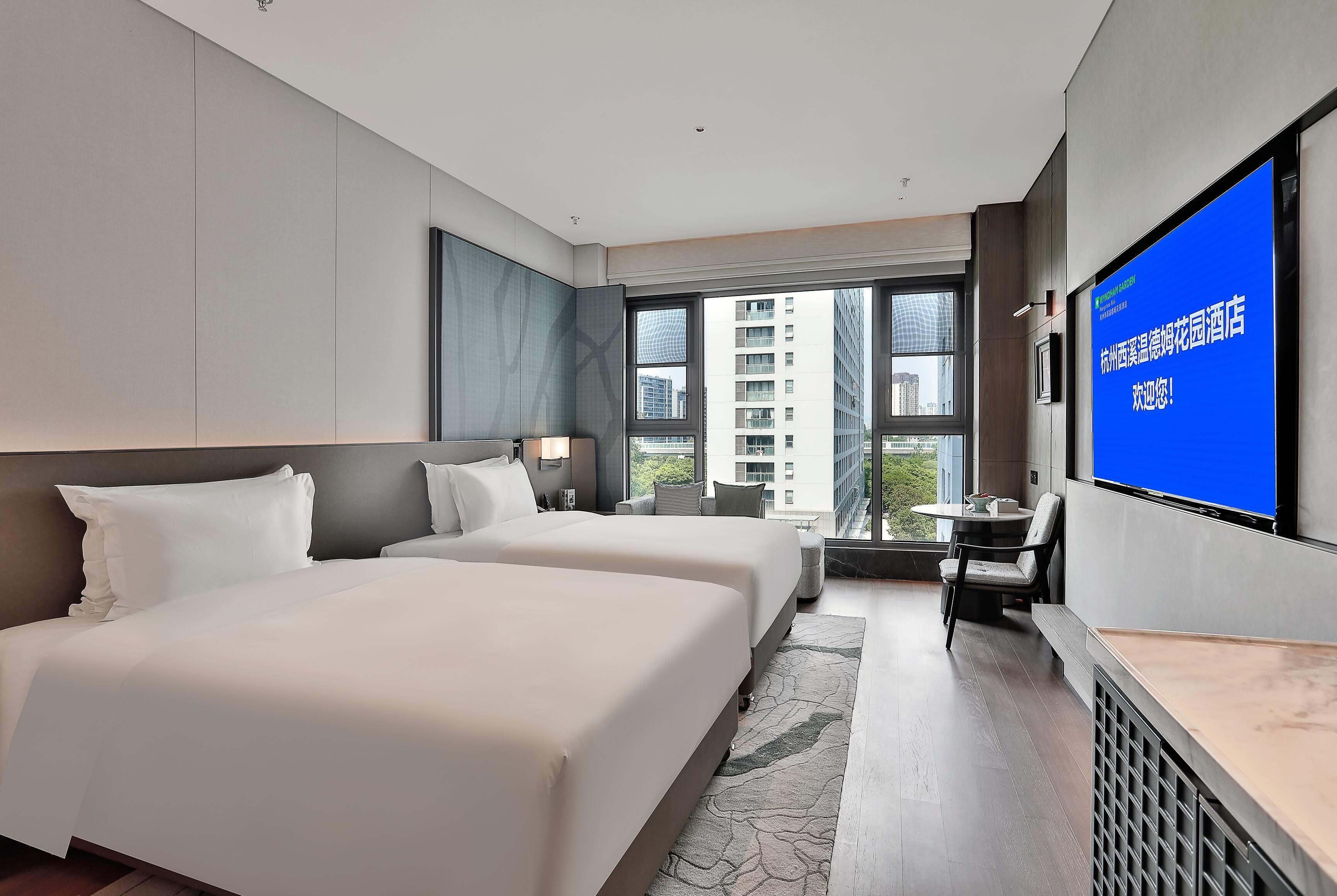 A hotel room with two beds, a desk, a chair, and a large TV screen.