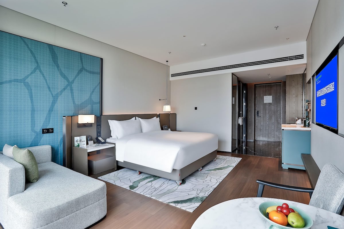 A hotel room with a bed, a desk, a chair, a TV, and a dining area with a table and fruit bowl.