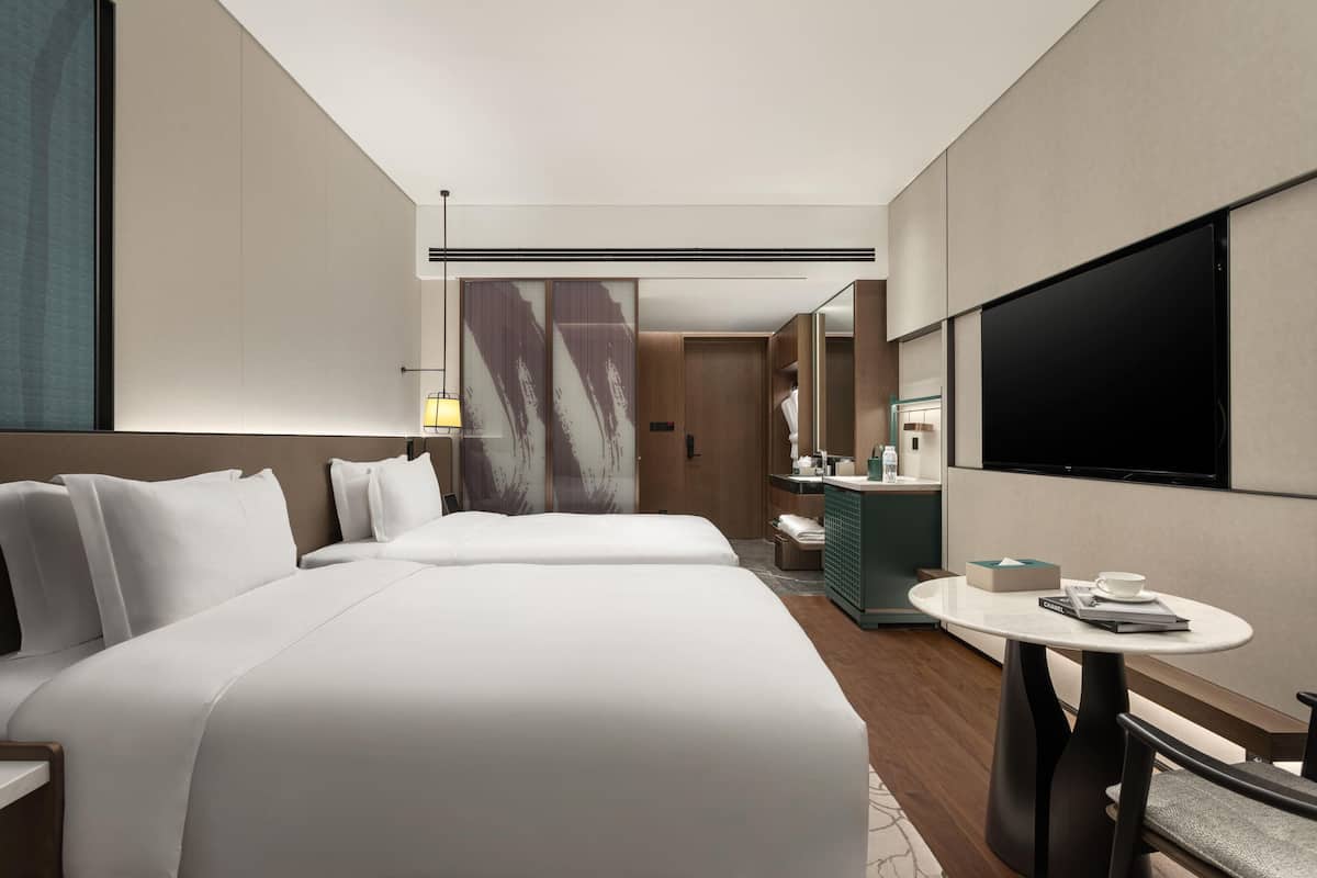 A hotel room with two beds, a TV, a small table with a cup and book, and a bathroom with a mirror and sink.