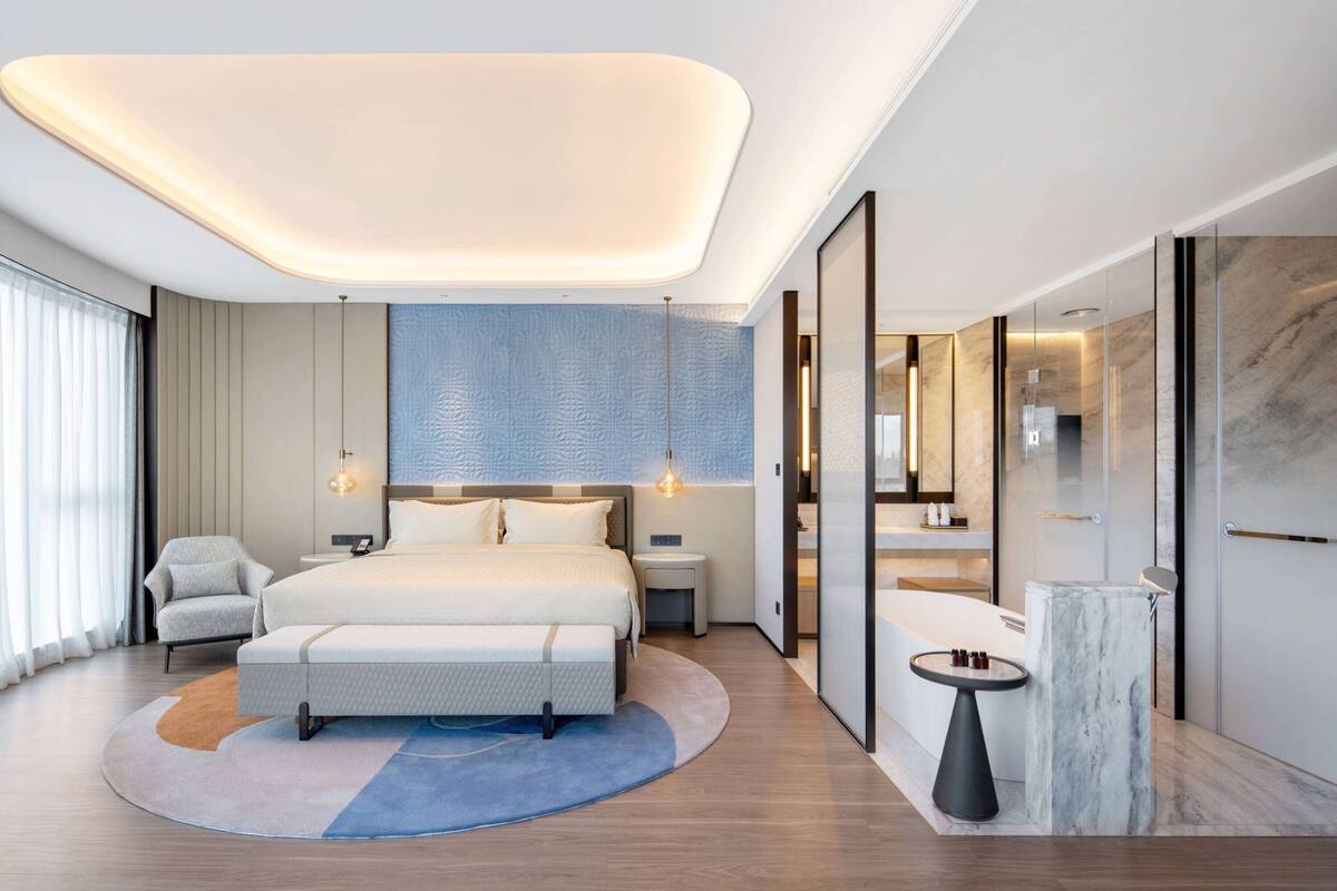 Una habitación de hotel moderna con una cama grande, mesitas de noche, un área de descanso y un baño con tocador de mármol.