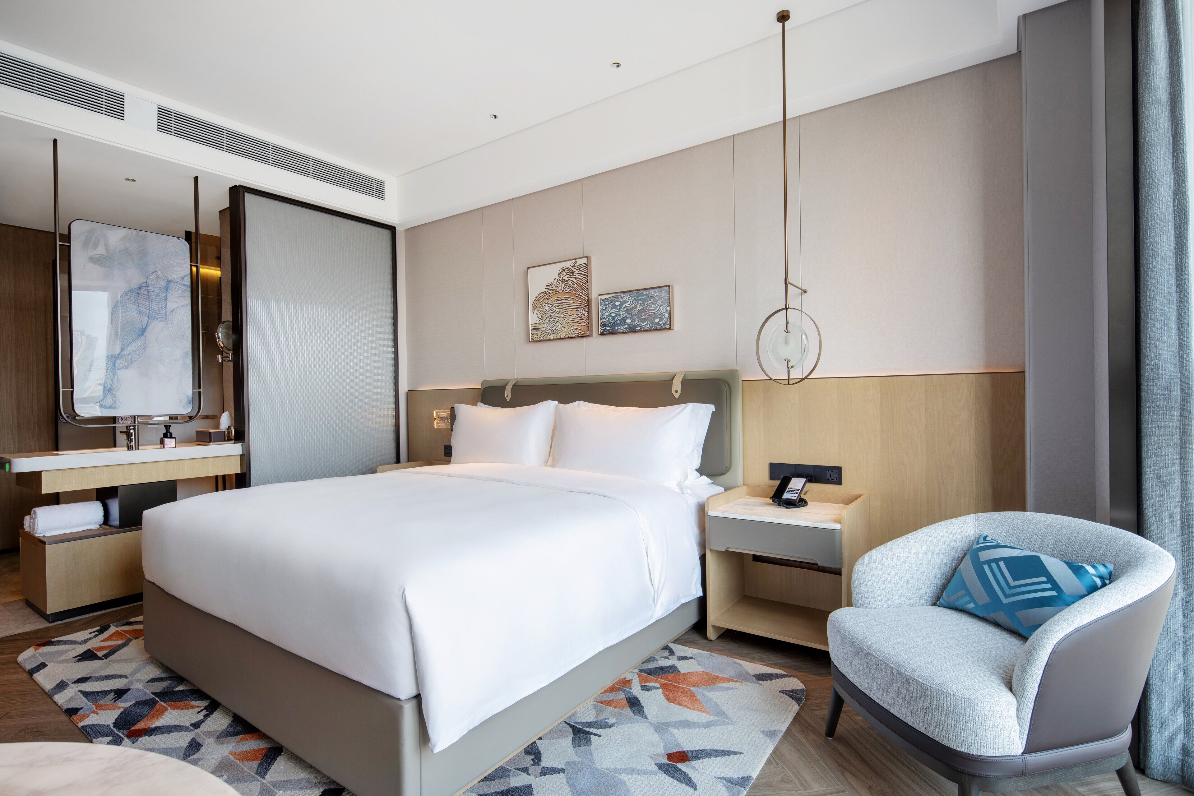 A modern hotel room with a large bed, a desk, a chair, and artwork on the wall.