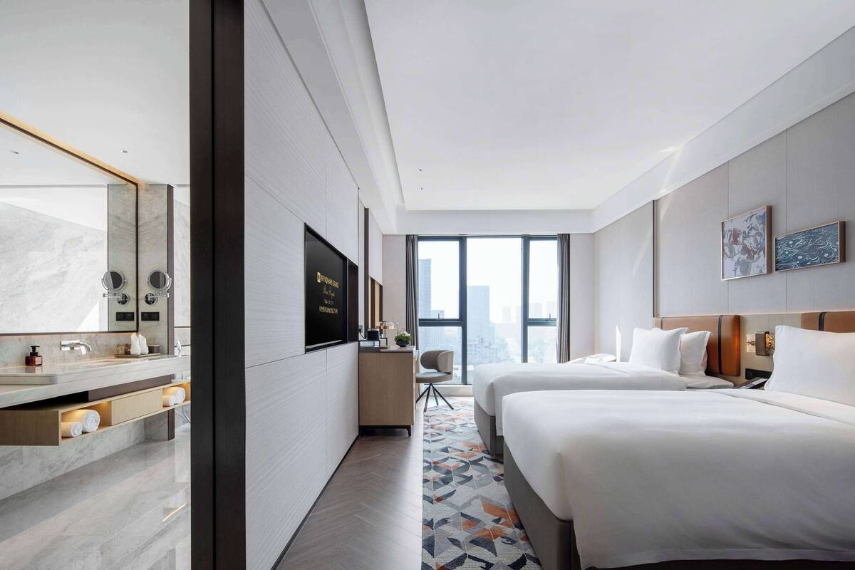 Habitación de hotel moderna con una cama grande, un escritorio con una computadora, un baño con espejo y vistas a la ciudad.