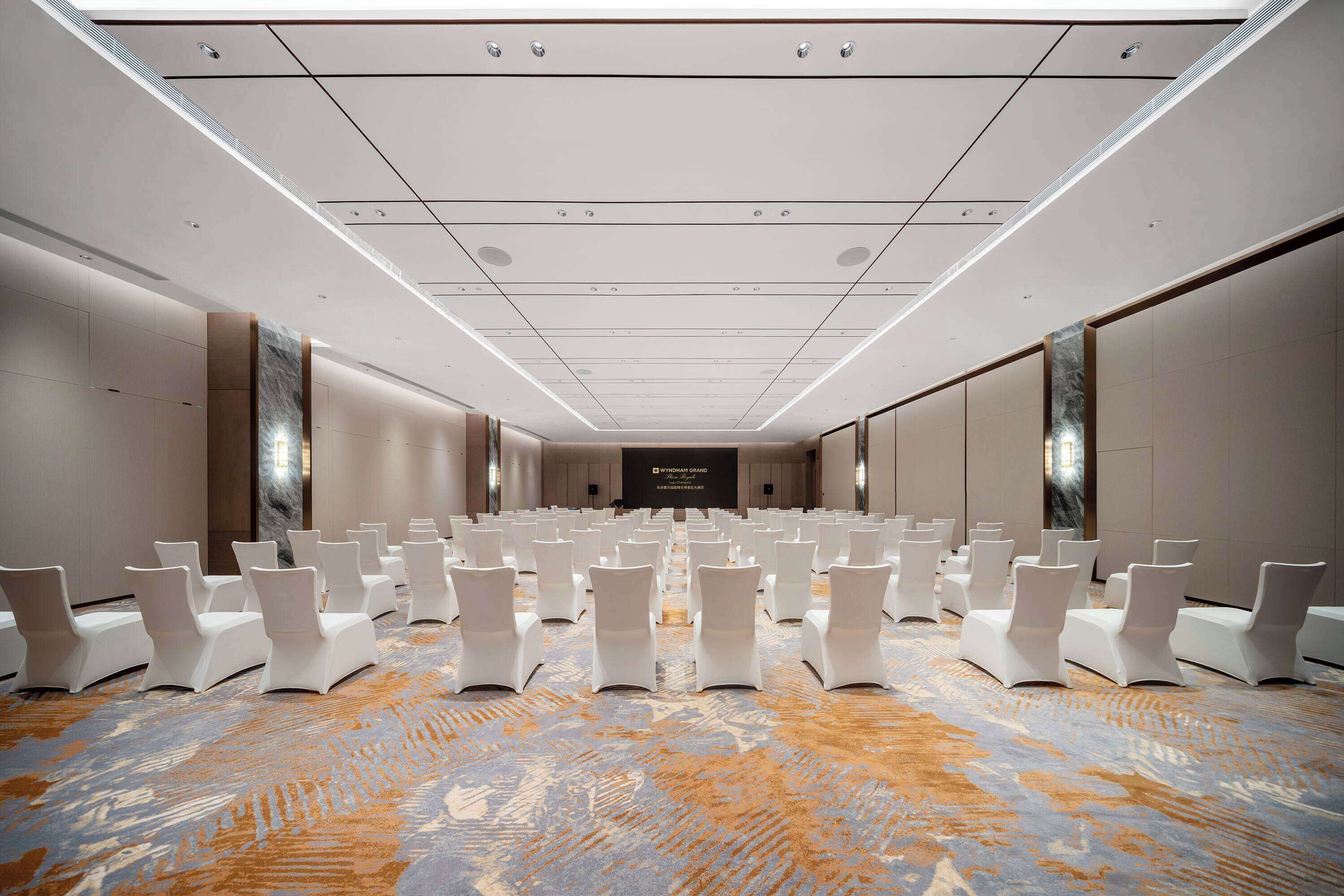 Un salón de conferencias con filas de sillas blancas, una alfombra estampada e iluminación empotrada en el techo.