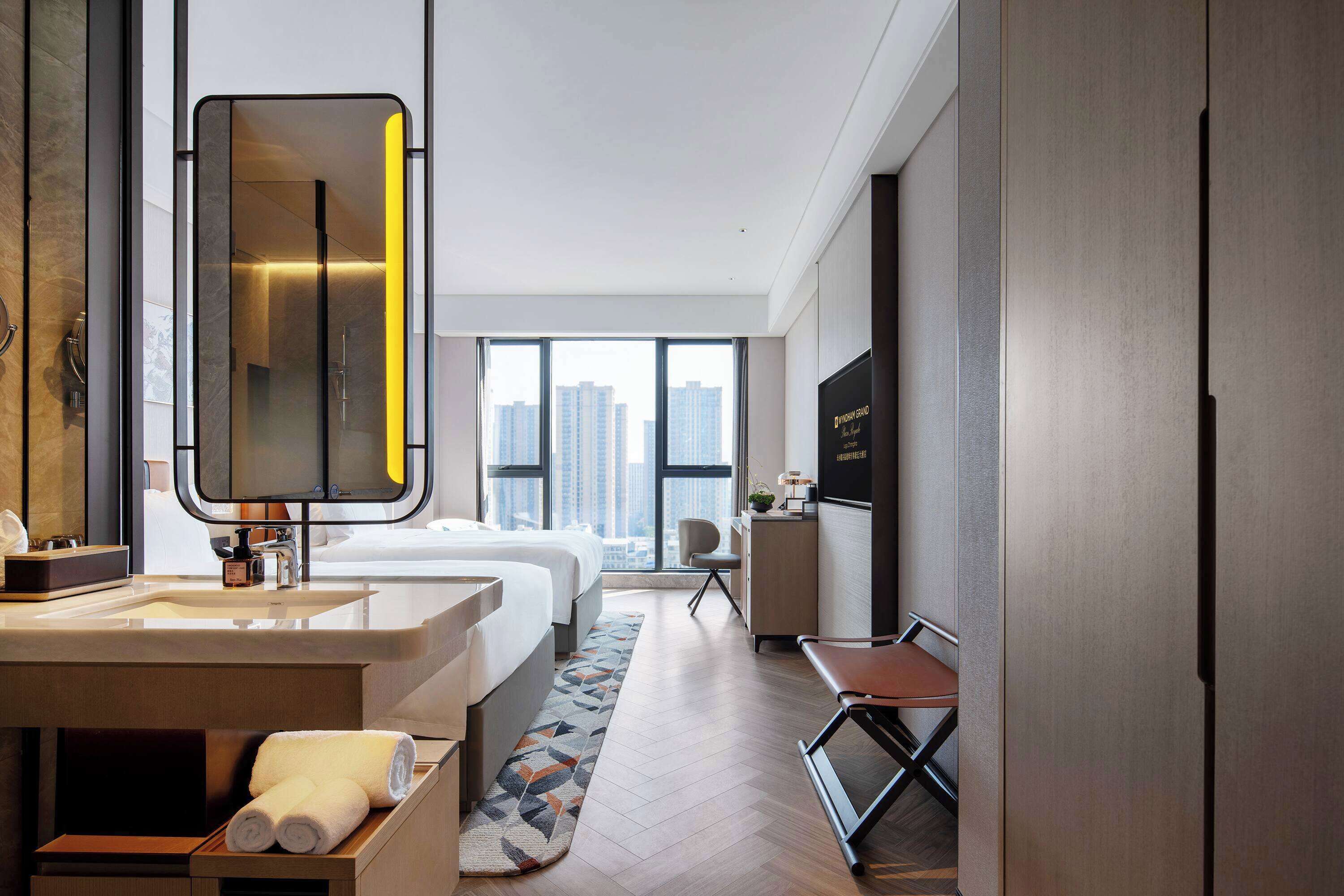 A modern hotel room with a large mirror, a bed, a desk, and a view of the cityscape through large windows.