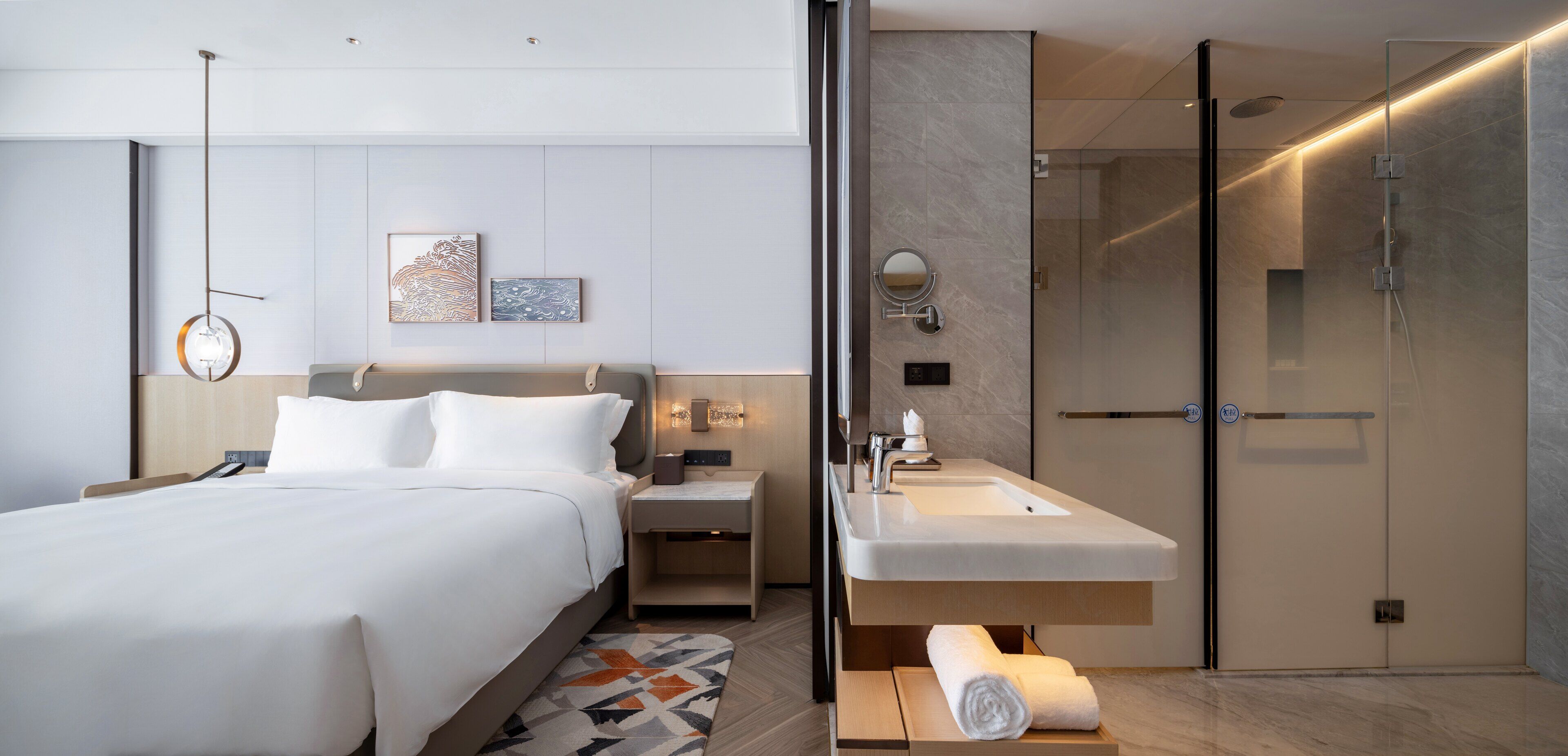 A modern hotel room with a large bed, a bedside table with a lamp, a bathroom with a sink and mirror, and a shower.