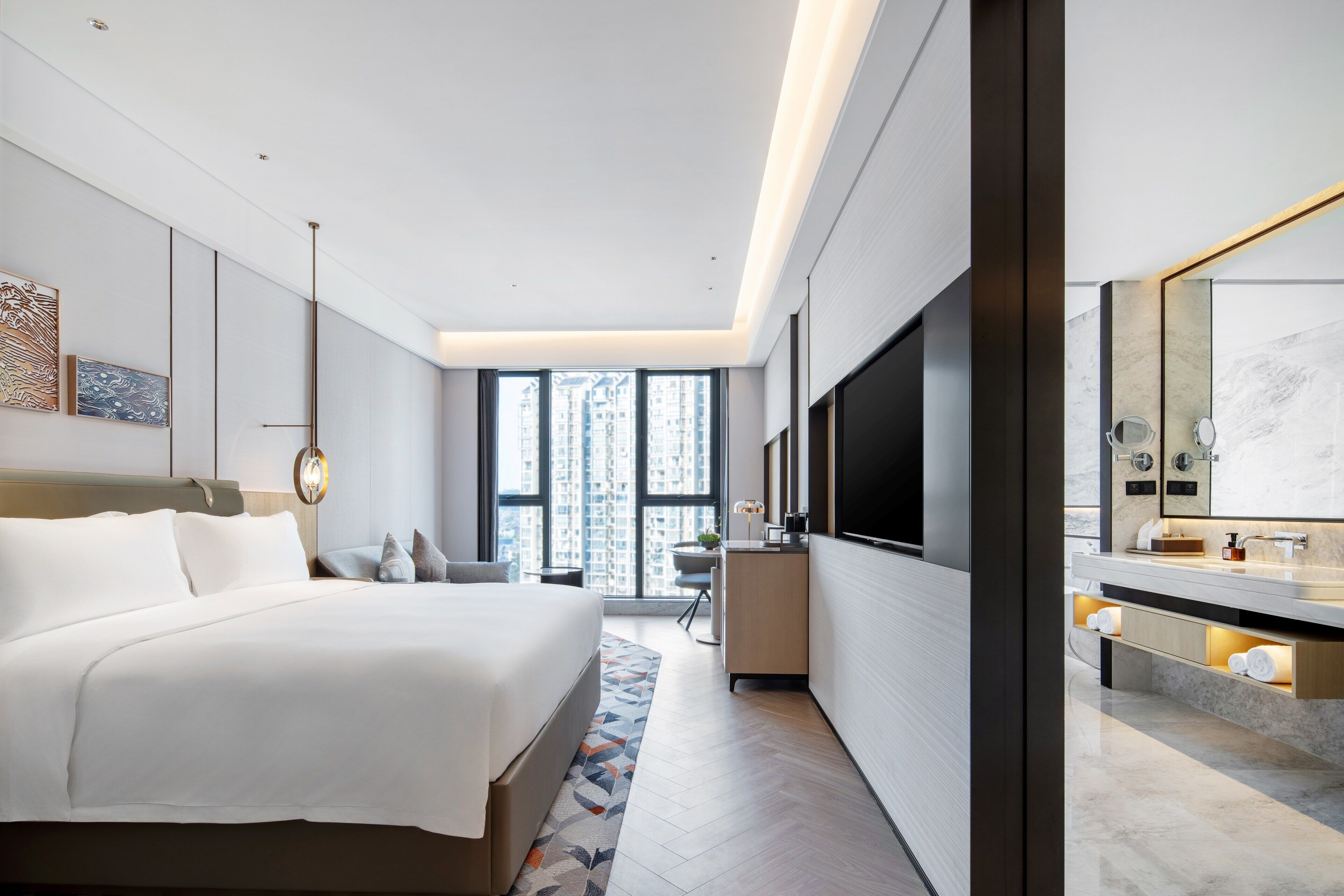 A modern hotel room with a large bed, a desk, a TV, and a bathroom with a mirror and sink.