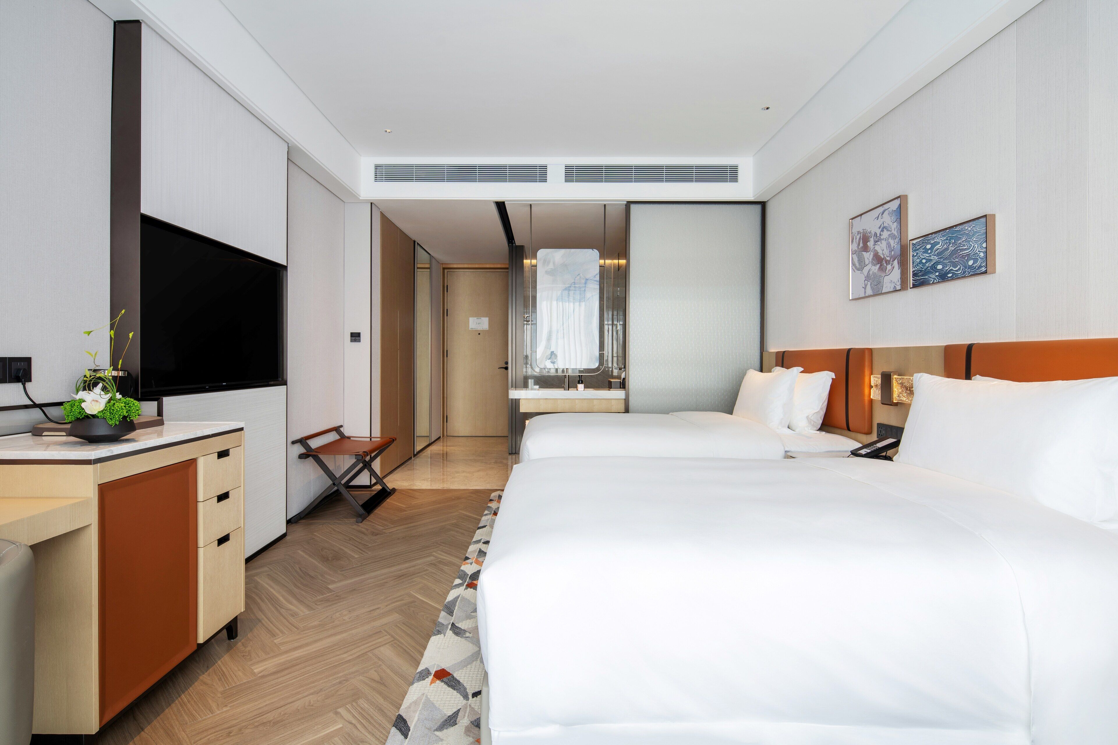 A modern hotel room with a large bed, a desk with a flat-screen TV, a small seating area, and a bathroom visible through a sliding door.