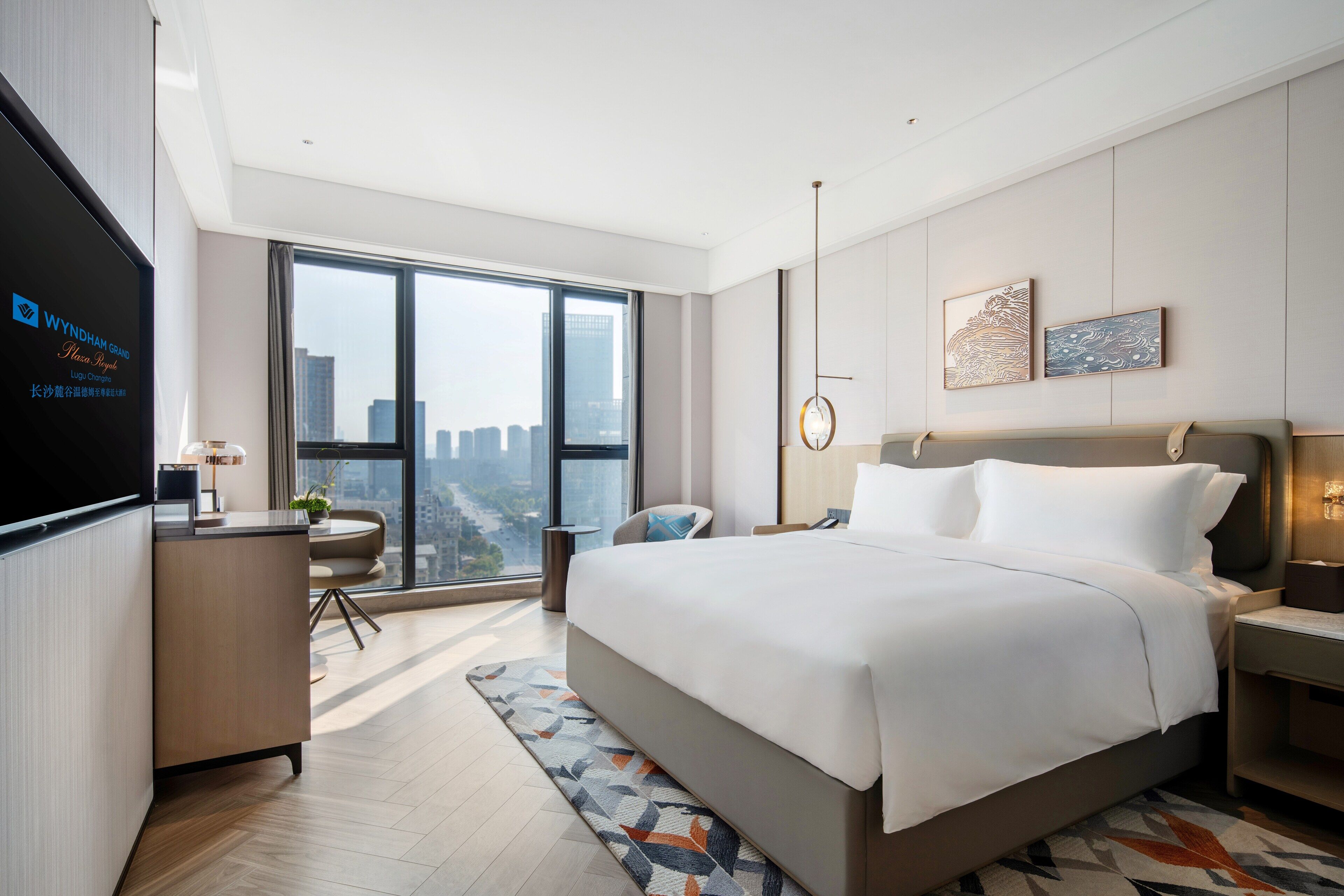 Ein modernes Hotelzimmer mit einem großen Bett, einem Schreibtisch, einem Stuhl und Blick auf die Stadt durch große Fenster.