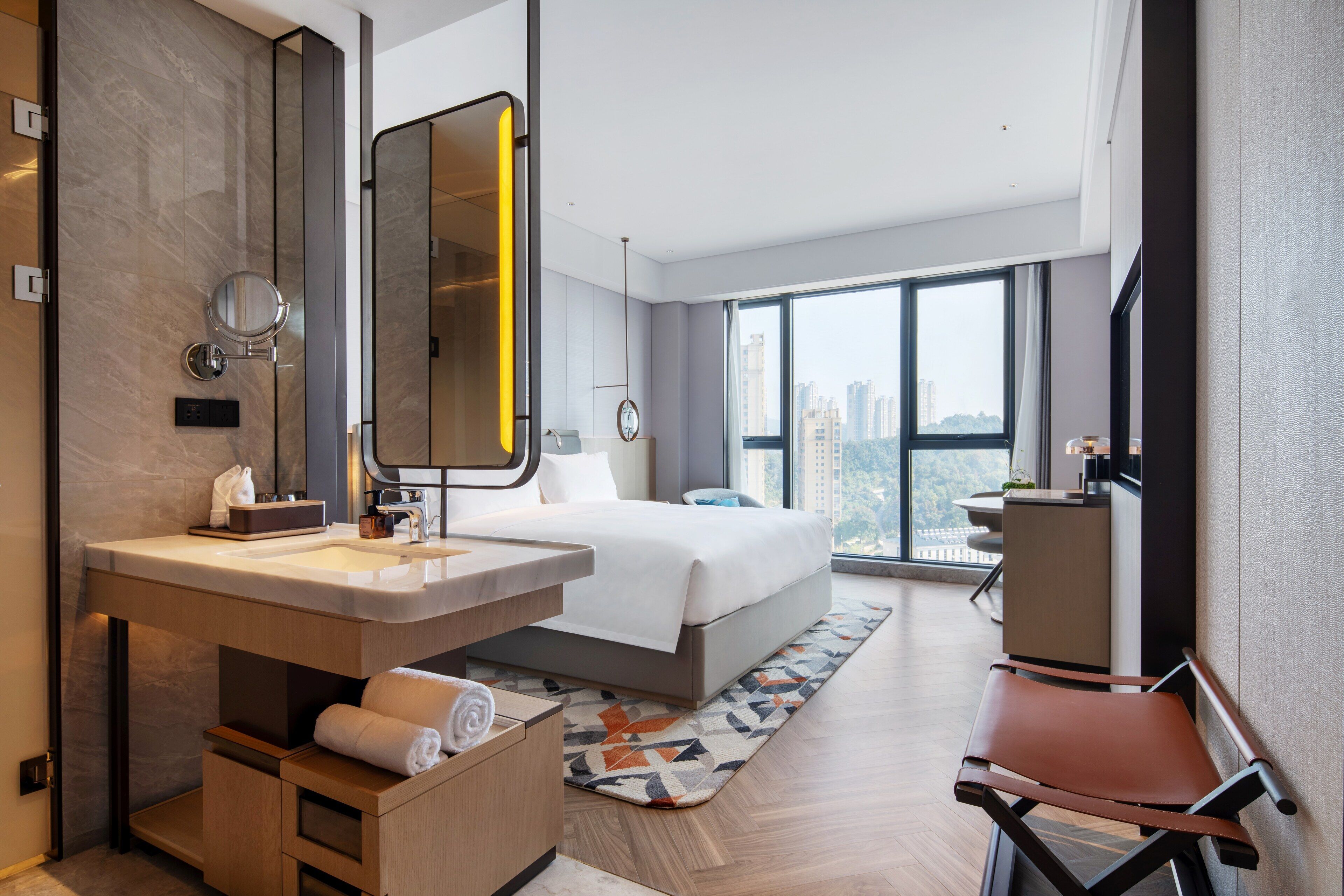 A modern hotel room with a large bed, a bathroom with a vanity, a desk, and a large window offering a city view.