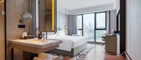 A modern hotel room with a large bed, a bathroom with a vanity, a desk, and a large window offering a city view.