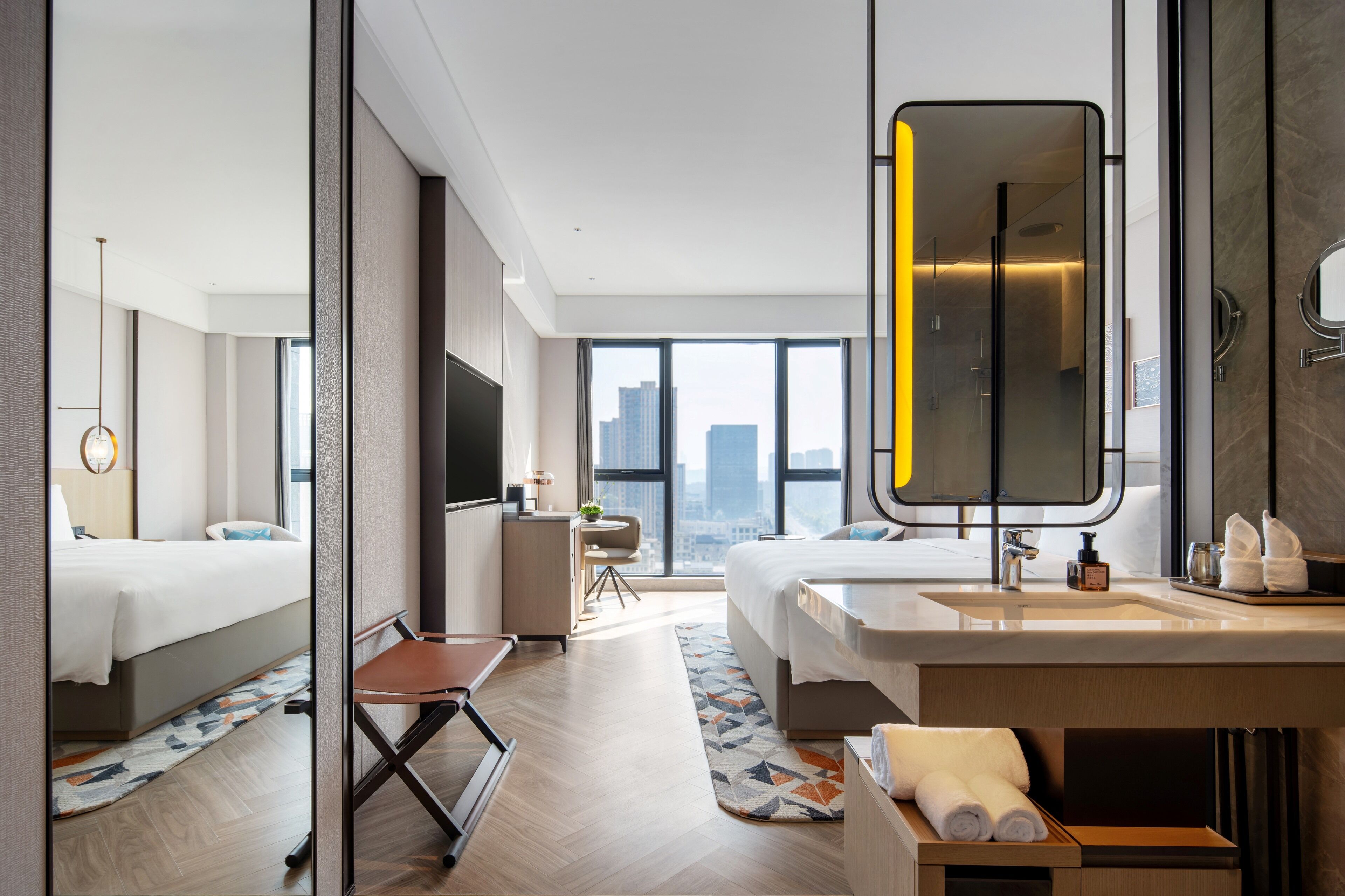 A modern hotel room with a large bed, a separate sitting area, a bathroom with a large mirror, and a city view.