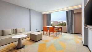 Una habitación de hotel moderna con sofá, una pequeña mesa redonda y un comedor con sillas. Hay un ventanal con cortinas y un televisor de pantalla plana montado en la pared.