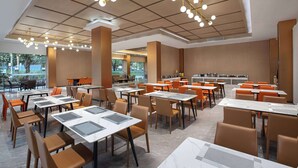 Un comedor moderno con varias mesas y sillas, una cocina al fondo y ventanas grandes que dejan entrar luz natural.
