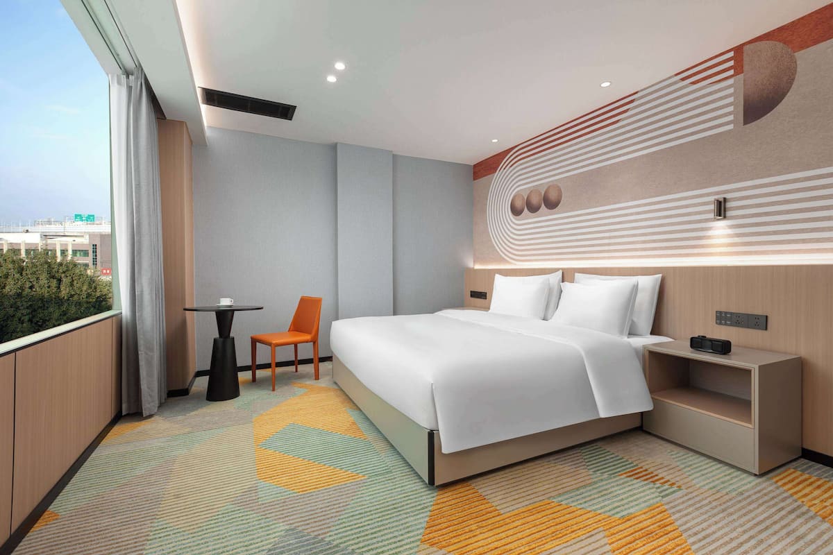 A modern hotel room with a large bed, a small table, a chair, and a view of the city.