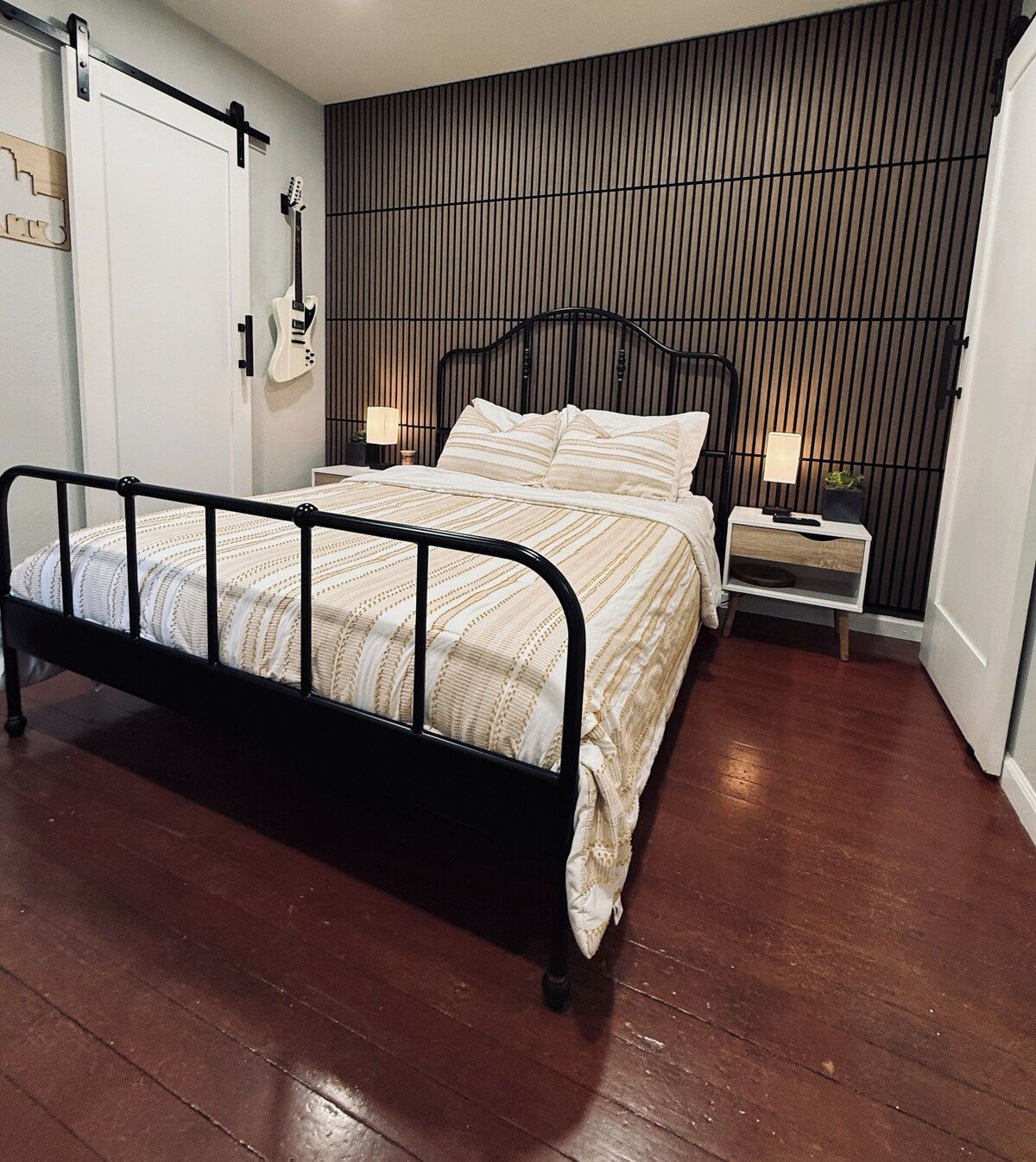 Un dormitorio con estructura de cama metálica negra, suelo de madera, una guitarra colgada en la pared y una mesita de noche con lámpara.