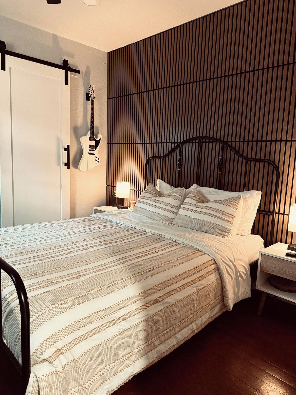 Un dormitorio con cama, mesita de noche, lámpara, guitarra y una pared con listones verticales.