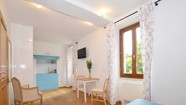 Habitación compacta con cama, una pequeña mesa de comedor, sillas, televisor y cocina con un mueble azul y una ventana con cortinas florales.