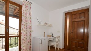 A small room with a wooden desk, a chair, a white refrigerator, a wooden door, and a window with a floral curtain. - Georgeous and Warm Living Downtown Rovinj - Standard Twin Room (Rovinj)
