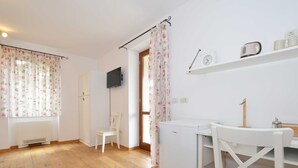 A room with wooden flooring, a white wardrobe, a small white chair, a white table, a white refrigerator, a flat-screen TV mounted on the wall, and a shelf with a clock and a kettle. - Georgeous and Warm Living Downtown Rovinj - Standard Twin Room (Rovinj)