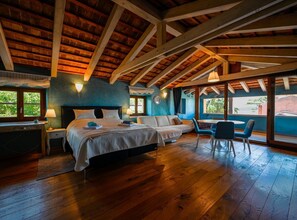 Une chambre spacieuse avec un grand lit, un canapé et un coin repas avec des chaises. La pièce est dotée de planchers en bois et d’un plafond en pente avec des poutres apparentes.