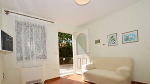 Une pièce avec un canapé beige, un radiateur blanc, une fenêtre ornée de rideaux en dentelle et une porte donnant sur l’extérieur.