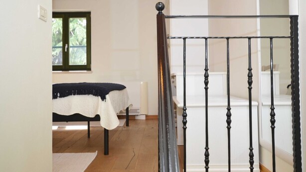 Une chambre à coucher avec un lit, un radiateur, une fenêtre et un escalier doté d’une rampe noire.