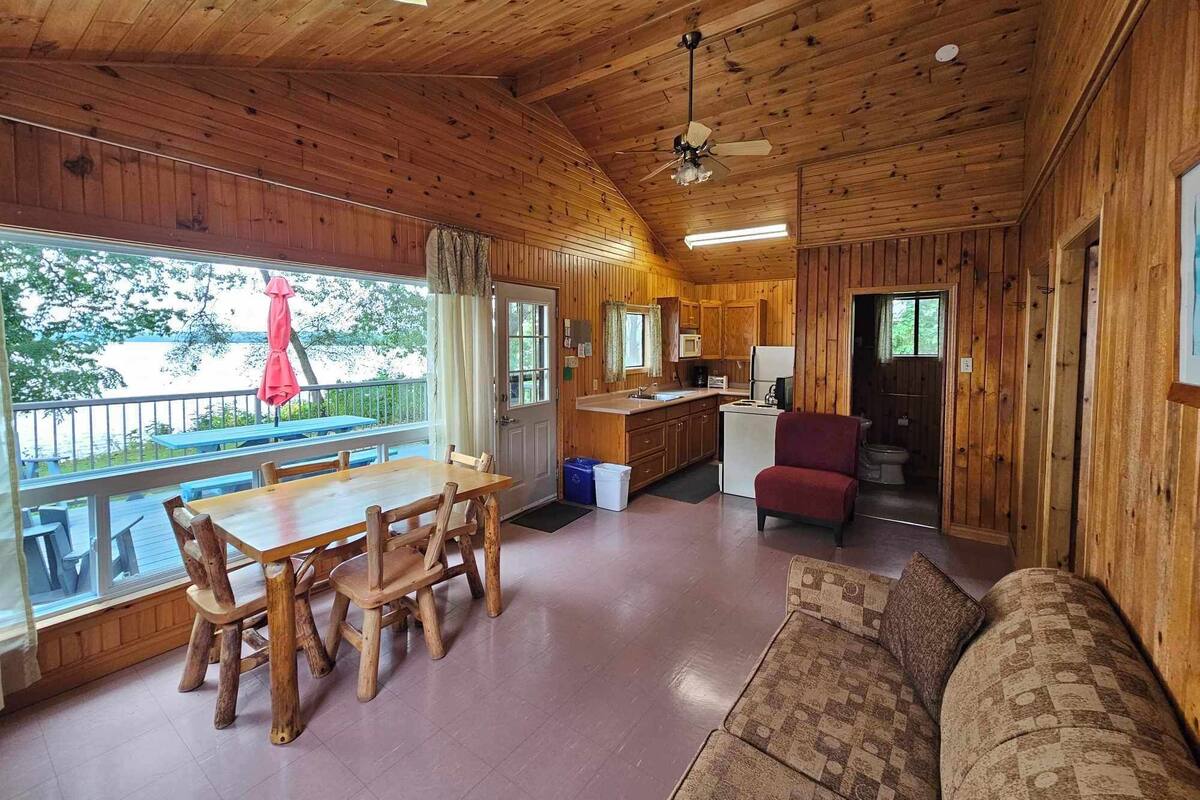 A cabin with a wooden interior, a dining area with a table and chairs, a kitchenette, a sofa, and a view of the outdoors.