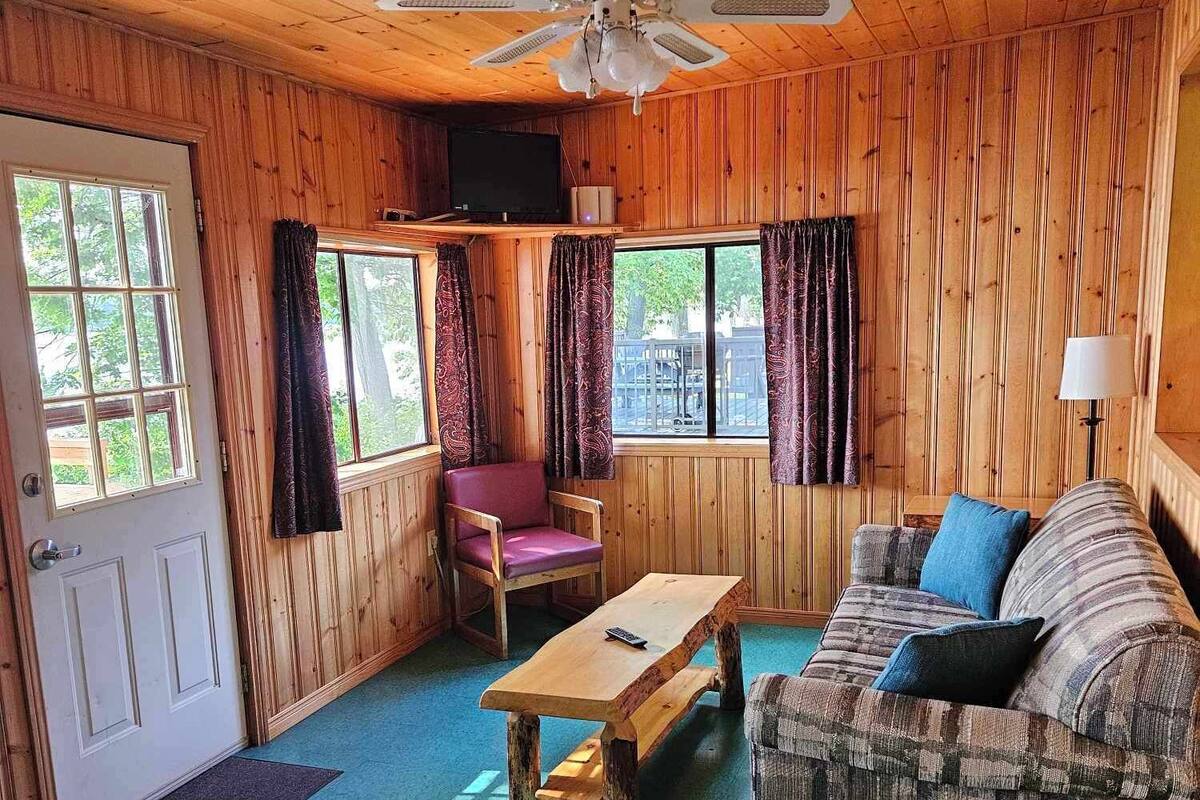 A cozy cabin interior with wooden walls, a ceiling fan, a sofa, a chair, a wooden table, and a television.