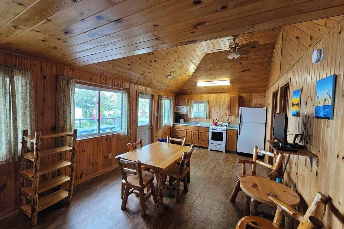 A cabin-style kitchen with wooden walls, a dining table, chairs, a stove, a refrigerator, and a television.