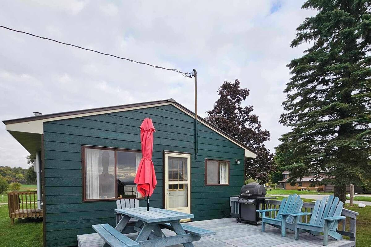 A small, green wooden cabin with a deck, picnic table, chairs, and a grill.