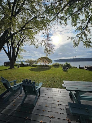 A lakeside picnic area with wooden benches and tables, a large tree, and a view of the lake and distant hills. - Southview Cottages Resort (Bailieboro)