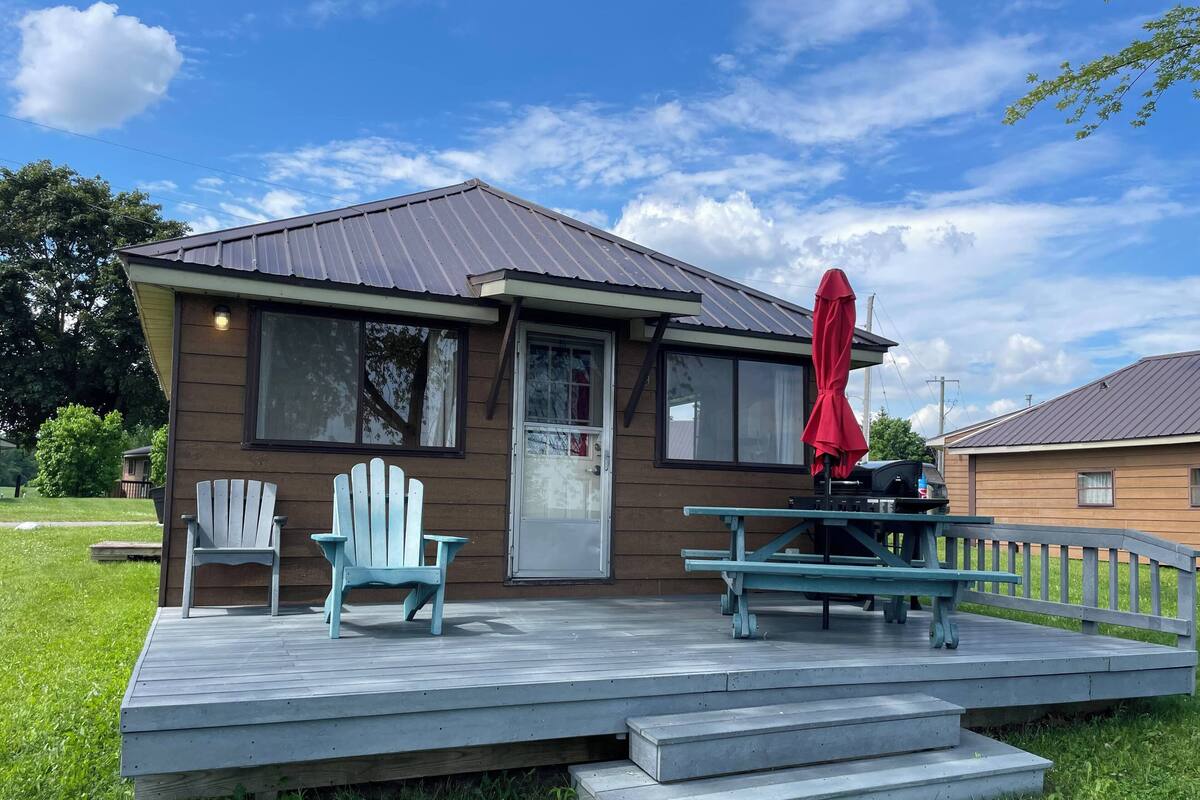 A small wooden cabin with a porch, two Adirondack chairs, a red umbrella, and a picnic table.