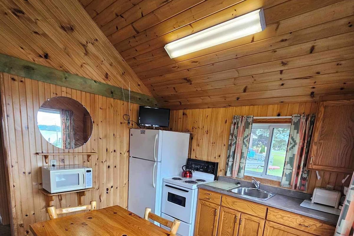 A wooden cabin interior with a dining table, chairs, a refrigerator, a microwave, a stove, and a sink.