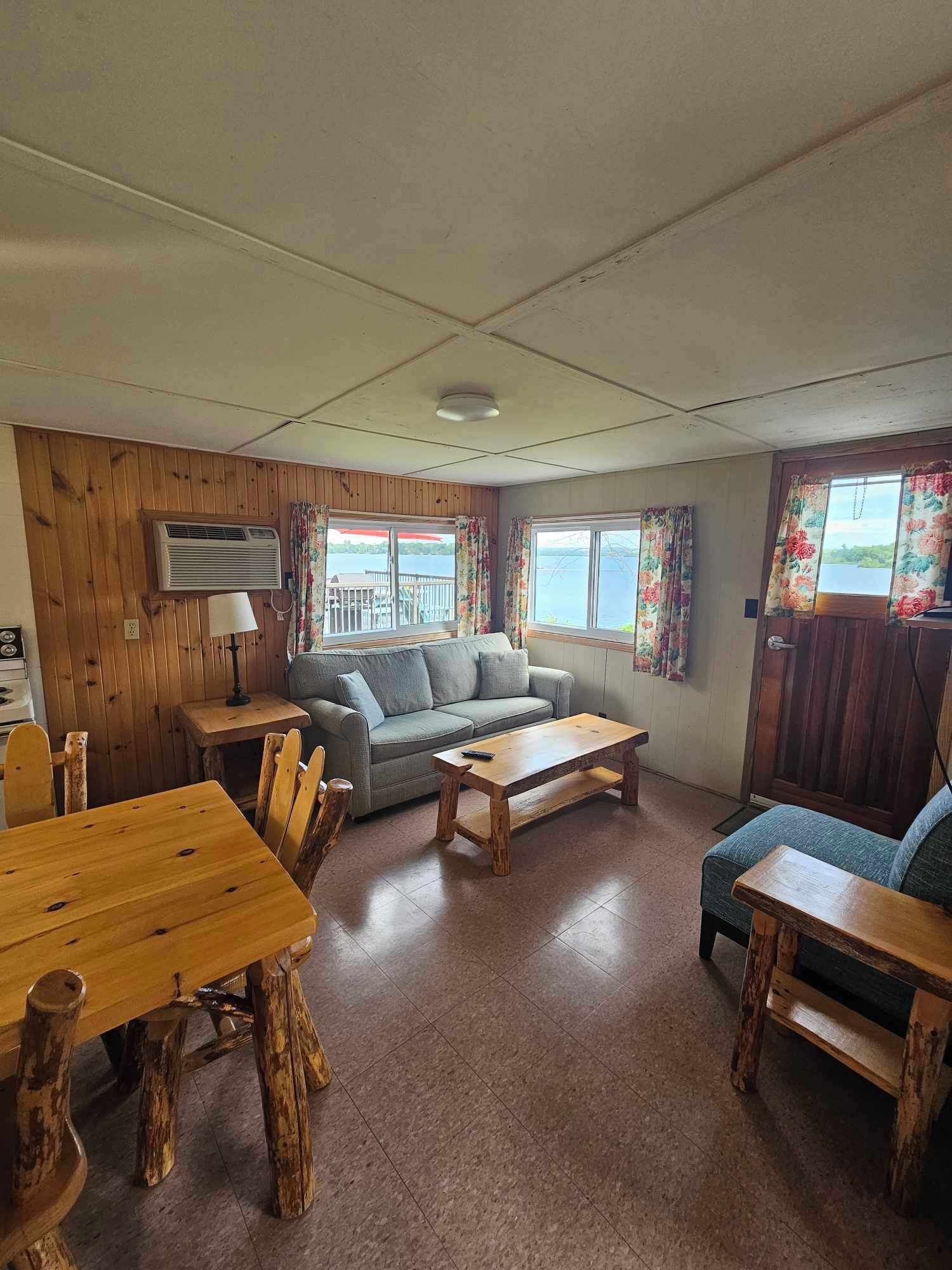 A cozy living room with a wooden table, a sofa, and a coffee table. There is a window with a view of a lake and a door with a wooden frame.