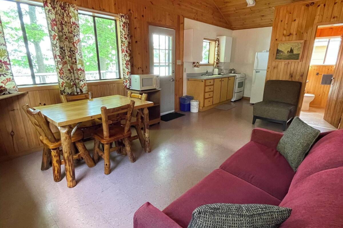 A wooden cabin interior with a dining table, chairs, a red sofa, a kitchen area, and a ceiling fan.