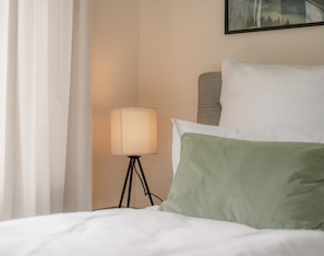 A bed with white bedding, a green pillow, a lamp on a tripod stand, and a painting on the wall.