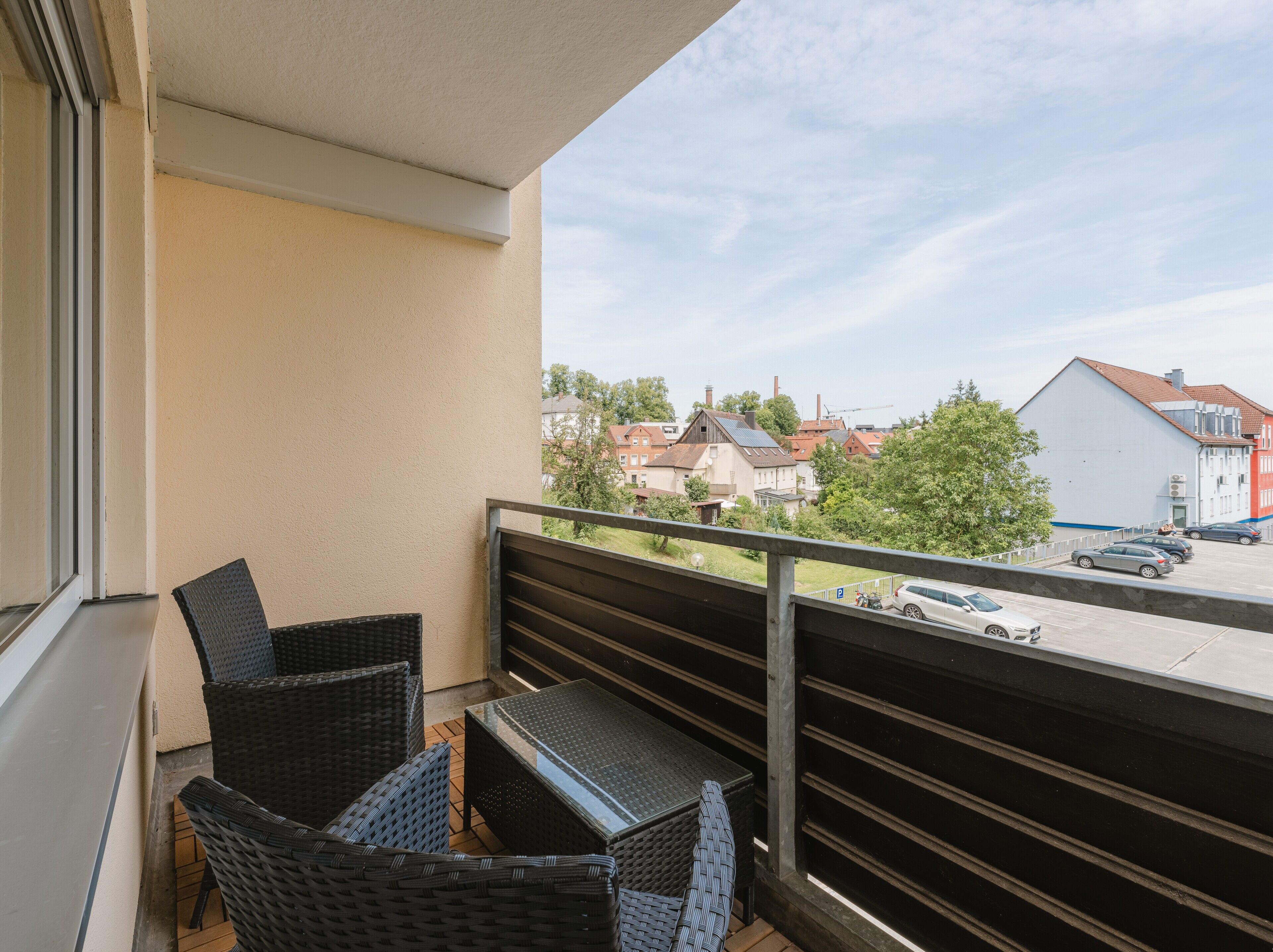 Ein Balkon mit Korbmöbeln, Blick auf eine Straße mit parkenden Autos und Gebäuden im Hintergrund.