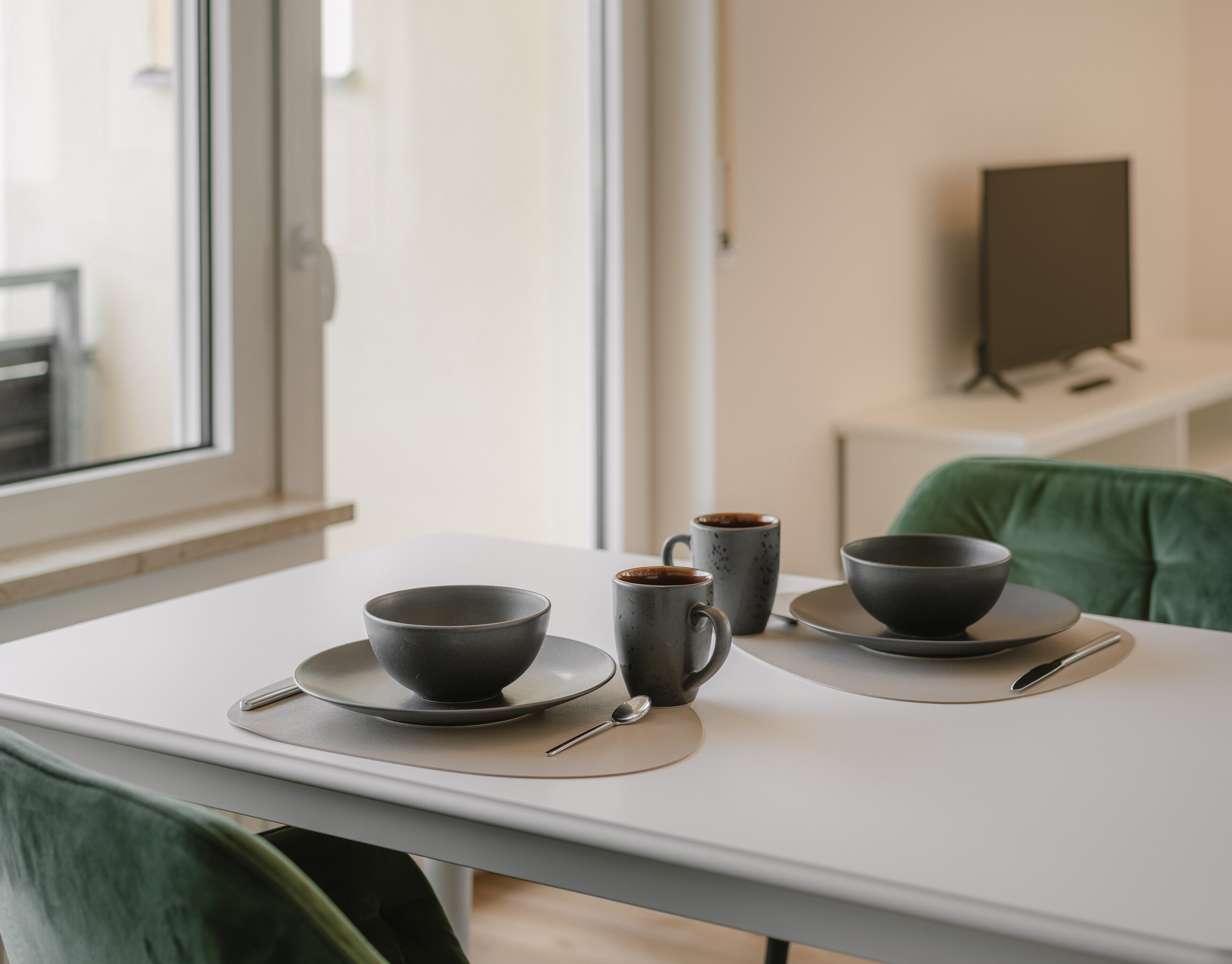 A minimalist dining table with two bowls and two cups, a spoon, and a green chair.