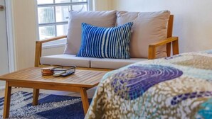 Um quarto com sofá, mesa pequena, colcha estampada, janela com persianas e um quadro na parede.