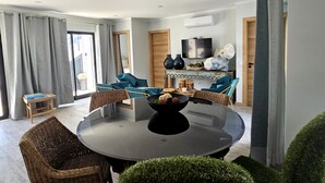 A modern living room with a round glass table, wicker chairs, a small side table, and a dining area with blue chairs.