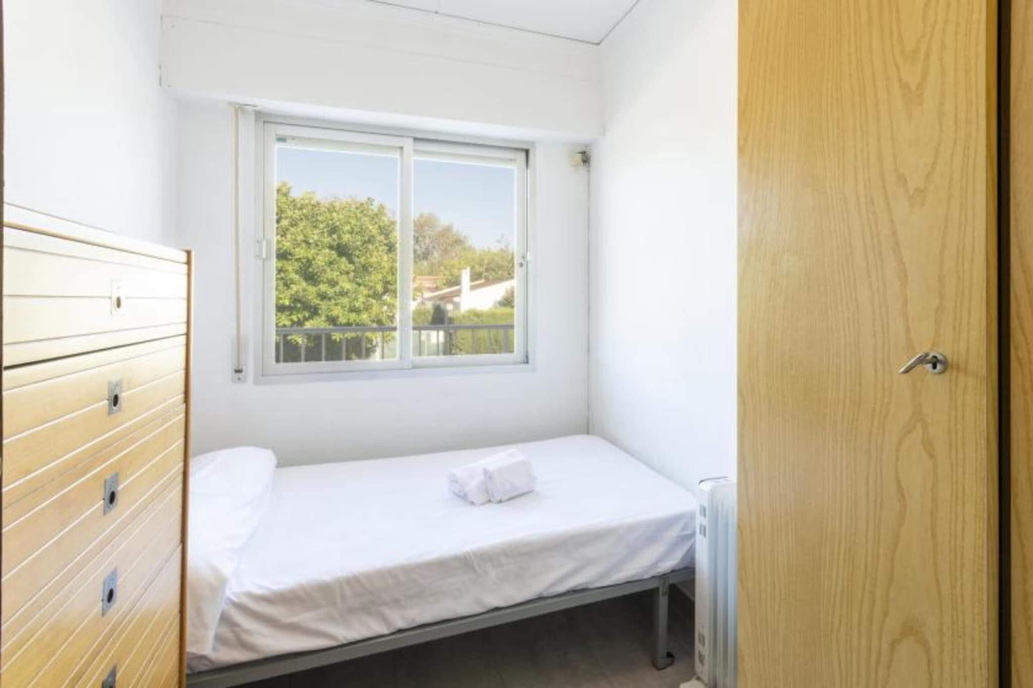 A small, single-bed room with a wooden dresser, a window with a view of greenery, and a wooden door.
