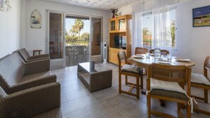 A living room with a sofa, a dining table with chairs, a TV on a wooden cabinet, and a sliding glass door leading to an outdoor area. - Cotton - Apartment In Urbanització Platja D'oliva (Playa De Oliva, Valencia)