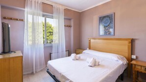 A hotel room with a bed, a bedside table, a window with sheer curtains, a radiator, and a painting on the wall. - Cotton - Apartment In Urbanització Platja D'oliva (Playa De Oliva, Valencia)