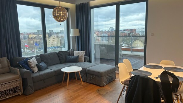 A modern living room with a grey sectional sofa, a round coffee table, and a dining area with a white table and yellow chairs. Large windows offer a city view. - Brabank 457 BestView by Grand Apartments (Gdansk)