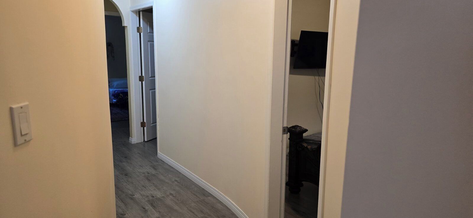 Ein schmaler Flur mit Holzboden, einer weißen Tür links und einem teilweise sichtbaren Zimmer mit Bett und Fernseher rechts.